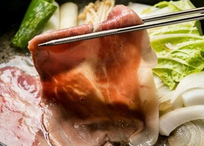 Two metal chopsticks picking up a thin slice of meat from the top of a shabu-shabu broth.